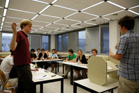 Sala donde aparecen un grupo de jovenes sentados en circulo, se encuentra dos jovenes de pie, uno, delante de un atril y otro, con la mano levantada para intervenir.