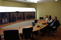 Tres pantallas en la pared, donde se reflejan personas dialogando con las que estan sentadas en la mesa de la la sala.