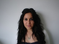 En la foto se ve el busto de una chica joven morena con el pelo largo y rizado mira de frente a la cámara.  Lleva una camiseta negra y una chaquetilla blanca.