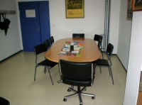 Imagen de una mesa de reuniones dentro de un despacho. Se aprecia una puerta al fondo de color azul, una mesa ovalada de madera y seis sillas negras rodeándola.