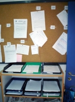 Se observa un corcho en una oficina lleno de documentos pinchados en él y unas bandejas de plástico debajo con distintos documentos.